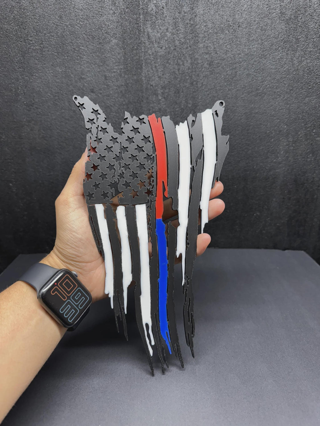 Tattered American flag wall decor with red and blue stripes and white accents, held in hand against a dark gray backdrop