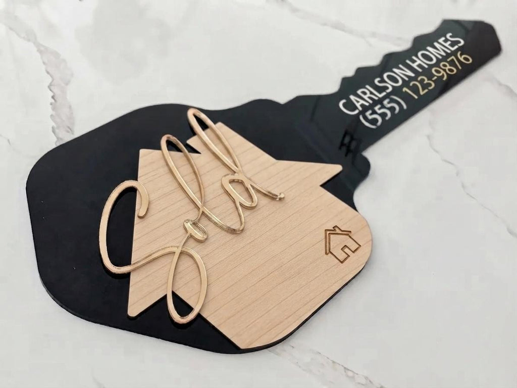 Black key-shaped Realtor Sold sign with wood-toned house, gold-tone script "Sold," and Carlson Homes text on marble