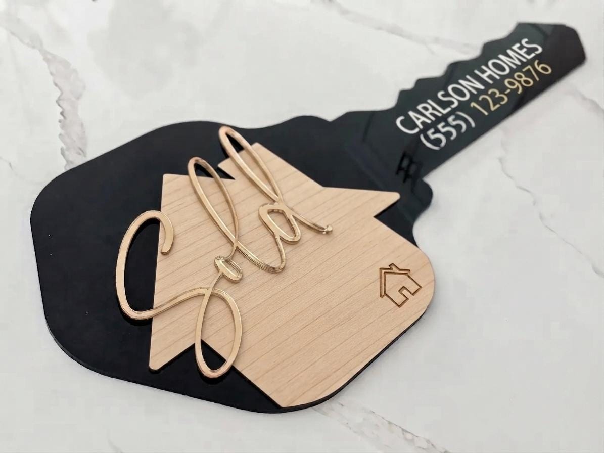 Black key-shaped Realtor Sold sign with wood-toned house, gold-tone script "Sold," and Carlson Homes text on marble