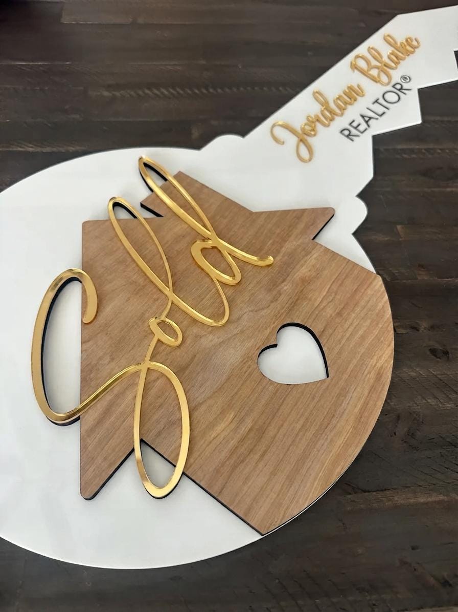 Key-shaped Realtor Sold sign with wood house, gold script "Sold" and "Jordan Blake REALTOR" text on dark wood surface